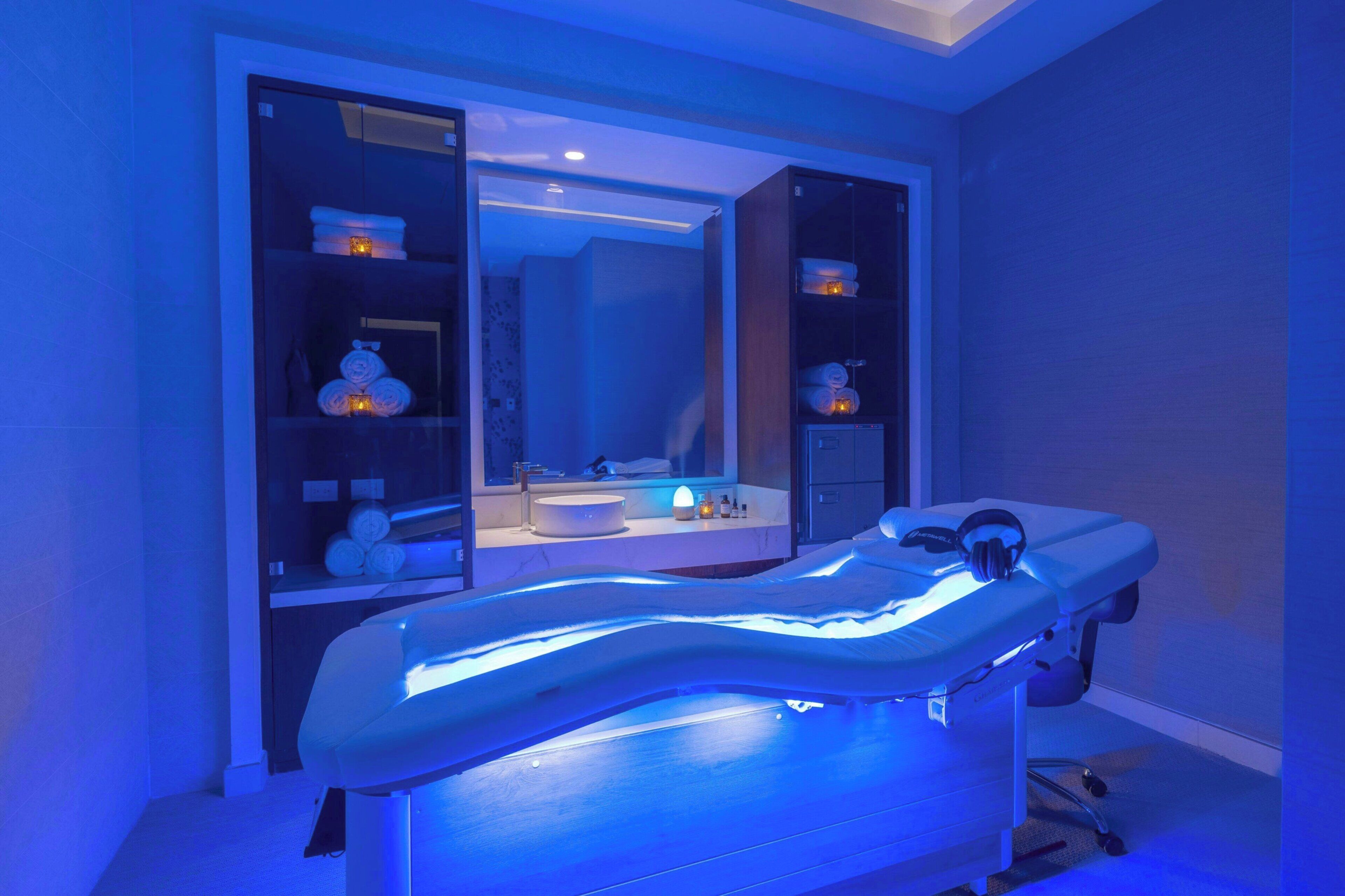 A medical examination table with a blue light, a mirror, and shelves with towels and candles.