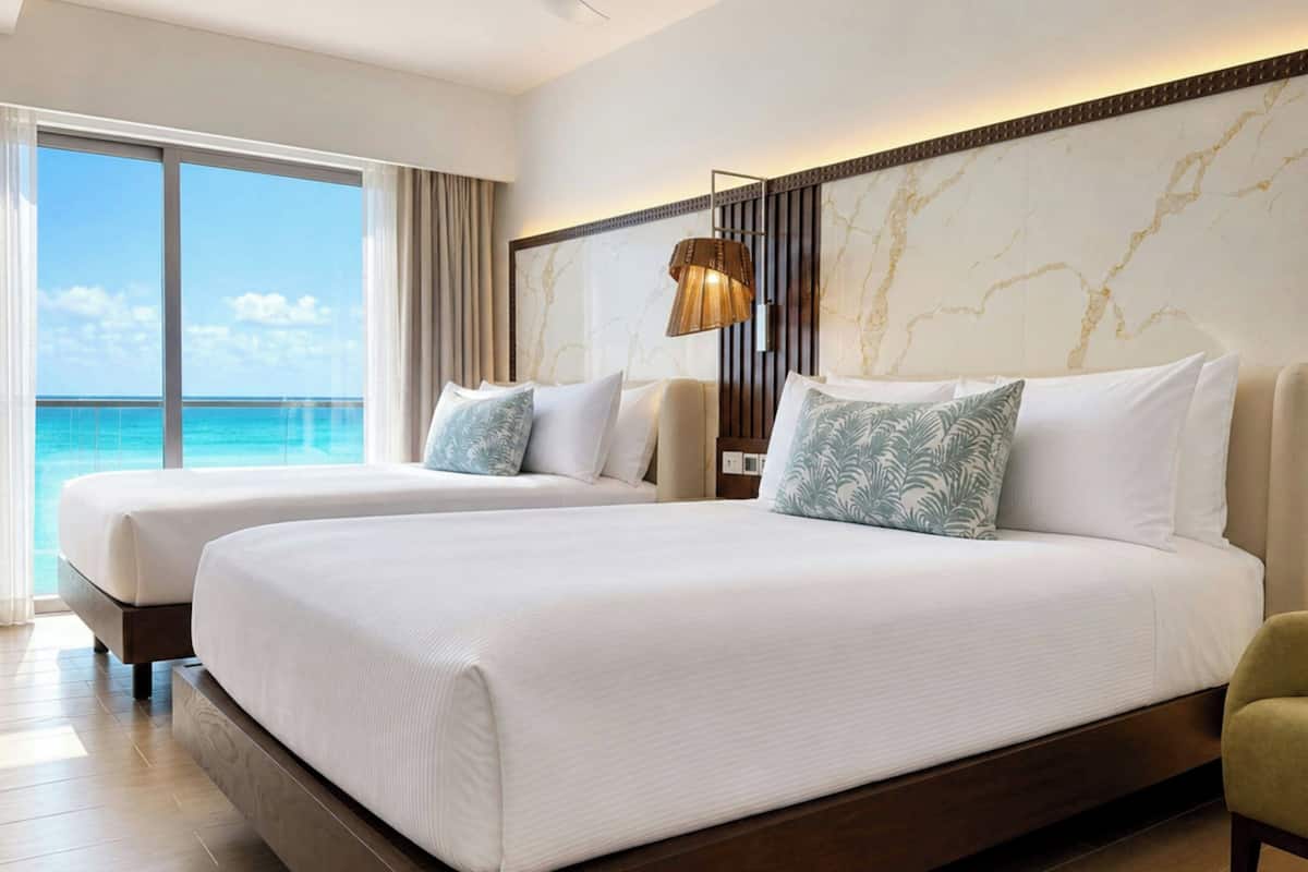 Hotelkamer met twee bedden, een groot raam met uitzicht op de oceaan, een marmeren wand en een moderne lamp.
