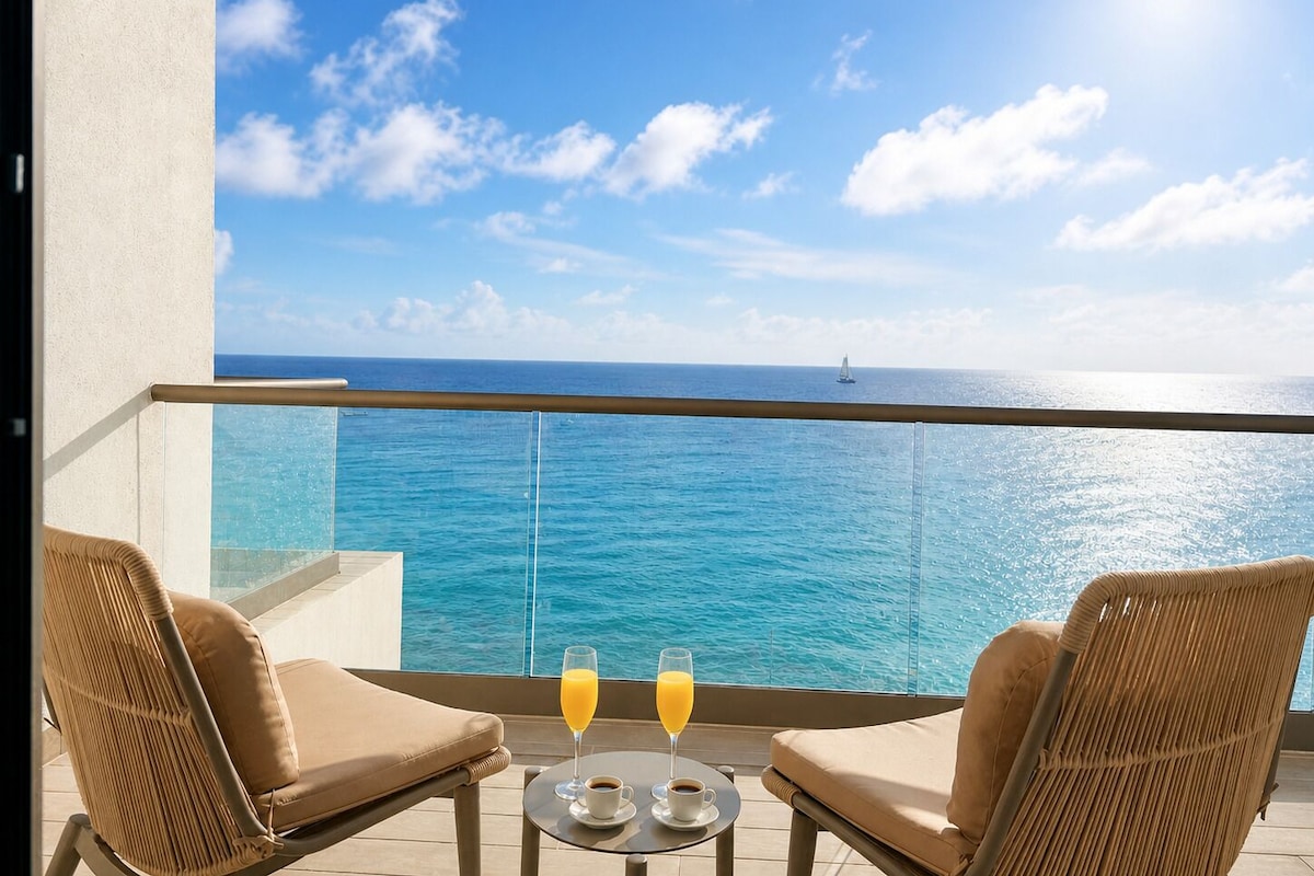 A balcony with two wicker chairs, a small table with two glasses of orange juice and a cup of coffee, and a view of the ocean.