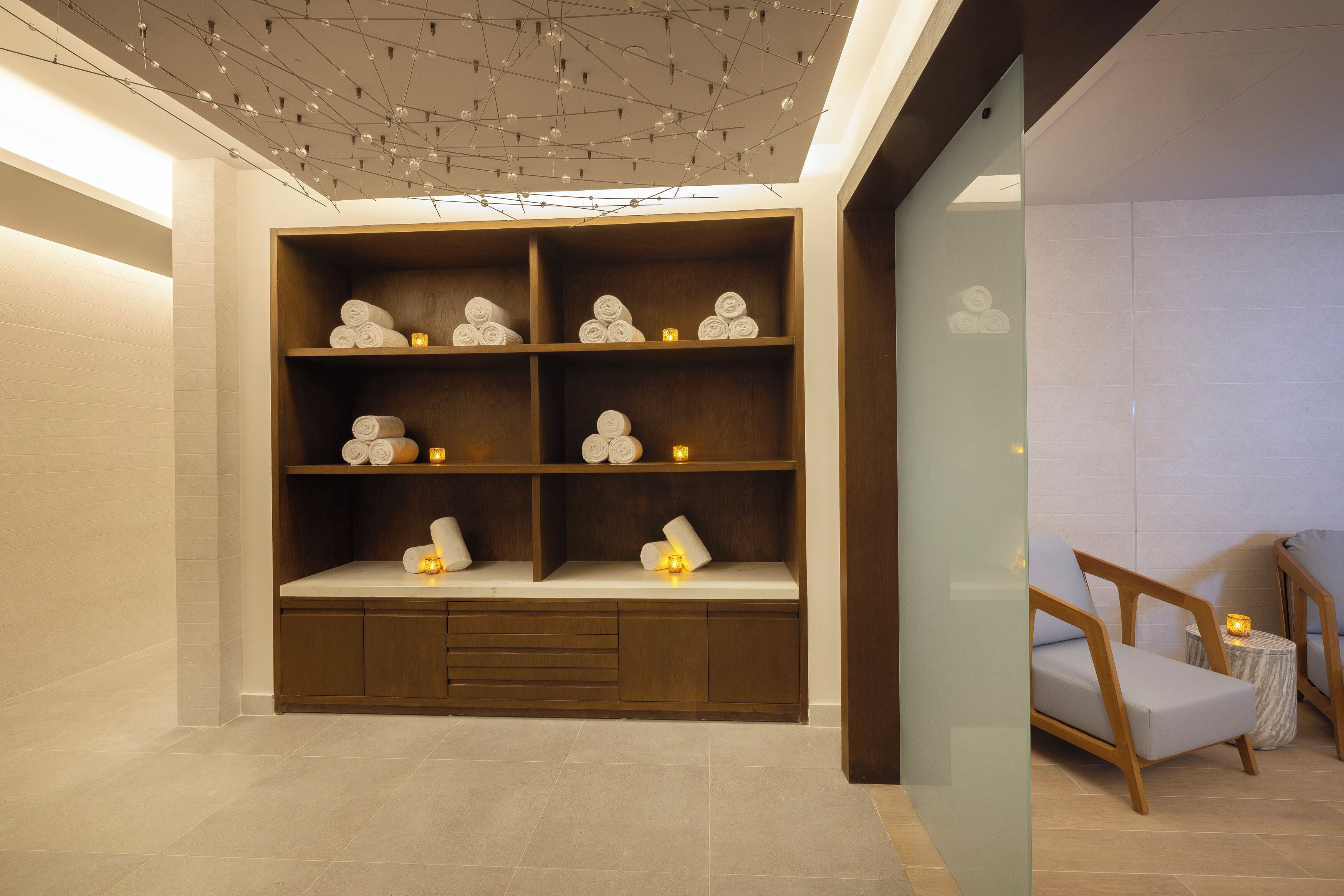 Een modern interieur met een houten plank waarop opgerolde handdoeken liggen, een klein rond tafeltje met een kaars en een fauteuil.