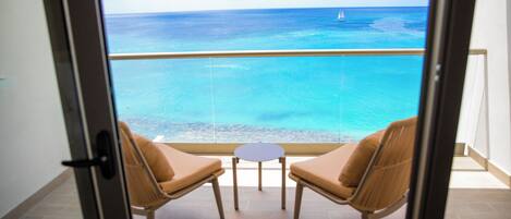 A balcony with two chairs and a small table, overlooking a clear blue sea with a sailboat.