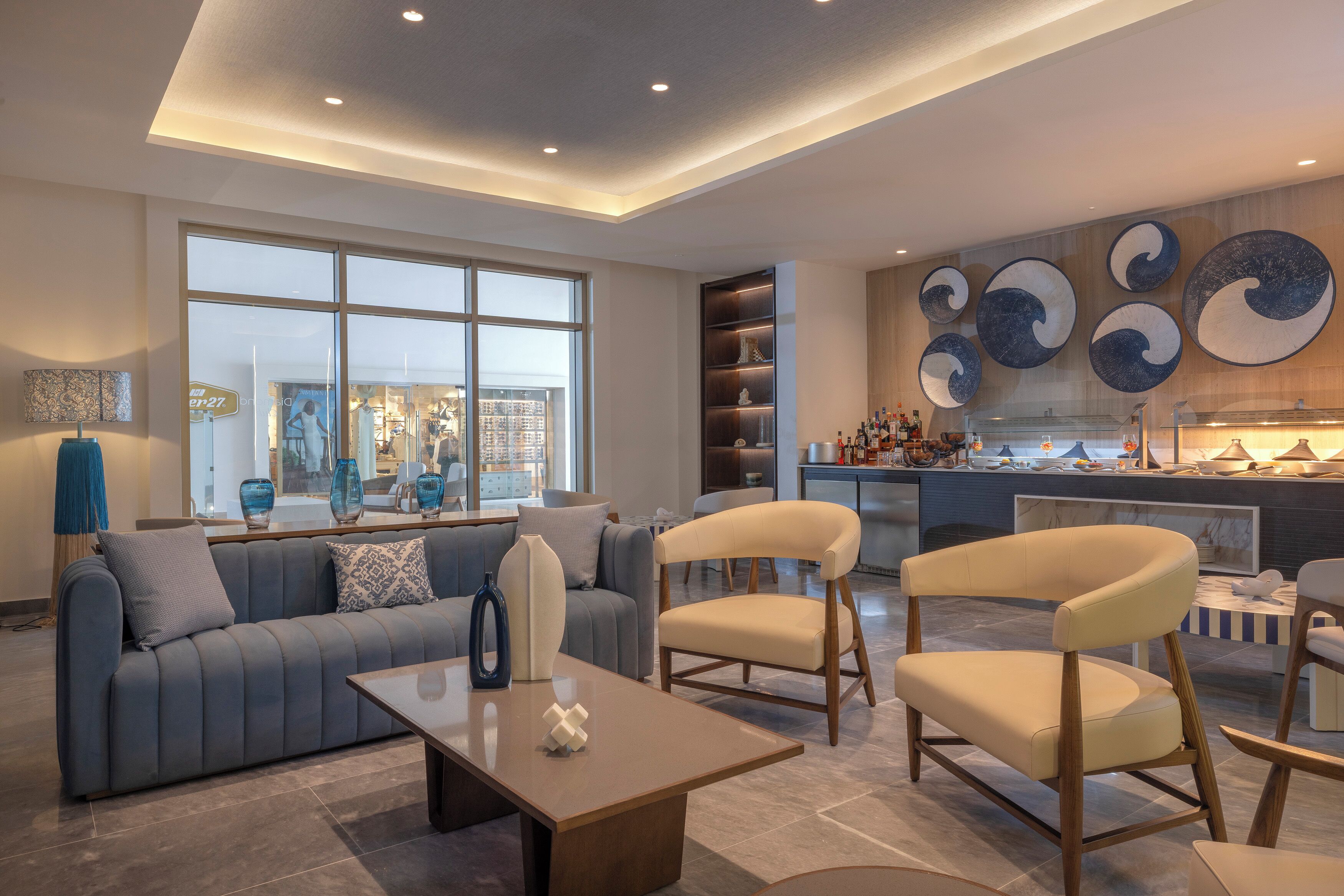 A modern living room with a sofa, chairs, a coffee table, and a kitchen area with a large window and decorative wall art.