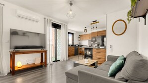 Un salon moderne avec un canapé, une table basse et un téléviseur posé sur une console en bois. Au fond, on aperçoit une cuisine avec des meubles en bois et un coin repas.