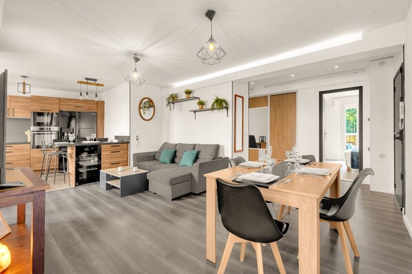 Cocina moderna con armarios de madera, horno empotrado y refrigerador. Cuenta con un comedor con mesa y sillas de madera, y un sofá gris en la sala.