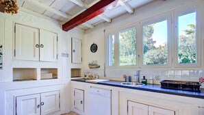 A kitchen with white cabinets, a dishwasher, a sink, and a window with a view of trees. - Catlove - Piscine Privée (Eus)