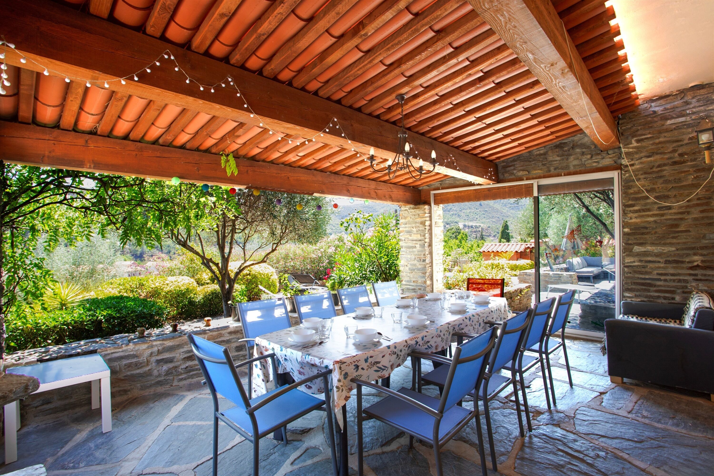 An outdoor dining area with a stone floor, wooden ceiling, and a view of a garden and mountains.