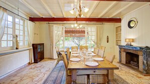 A dining room with a wooden table set for four, surrounded by wicker chairs, a chandelier, and a fireplace. - Catlove - Piscine Privée (Eus)