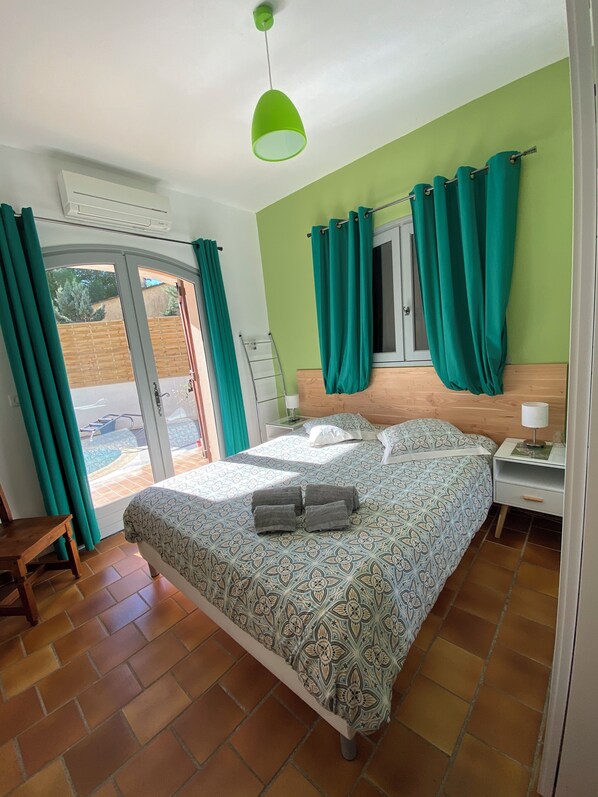 A bedroom with a bed, a nightstand, a lamp, and a view of the outside through a sliding glass door. - Maison d'hôtes naturiste Villa PaulAna (Cap d'Agde)