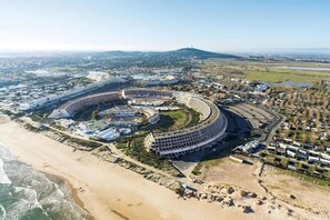 A large, circular building complex with a beachside location, surrounded by residential areas and a hilly landscape in the background. - Maison d'hôtes naturiste Villa PaulAna (Cap d'Agde)