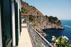 A coastal property with a balcony overlooking the sea, a rocky hillside, and a clear sky.