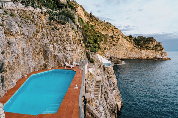 A cliffside swimming pool with steps leading down to the water, surrounded by rocky terrain and overlooking the sea.