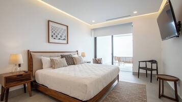 Un dormitorio amplio con una cama grande, una mesita de noche con lámpara, una obra de arte enmarcada y un televisor montado en la pared.