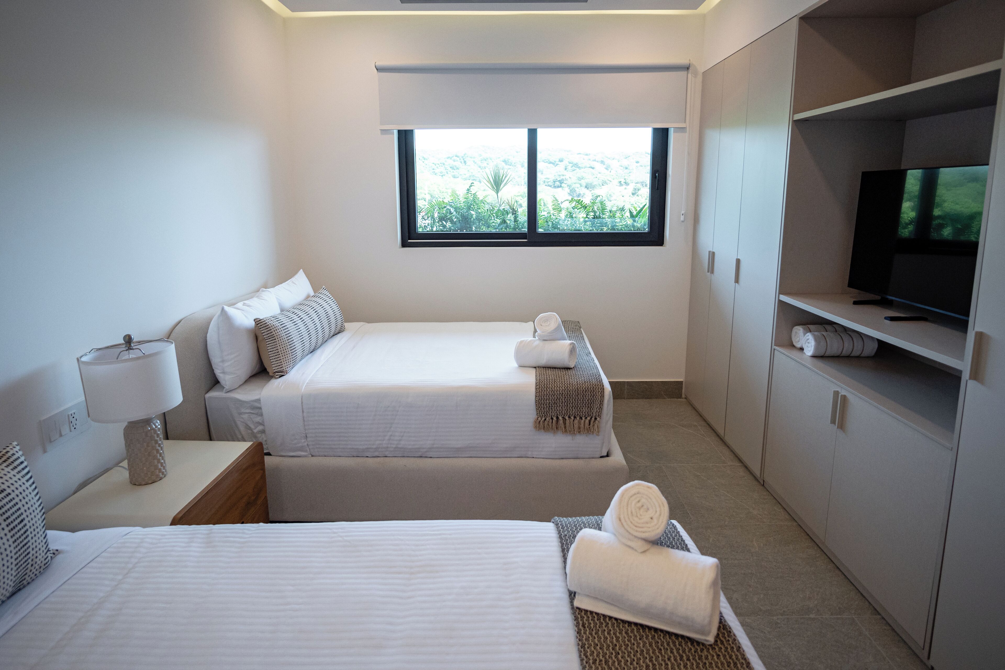 Quarto de hotel moderno com duas camas, televisão e uma janela grande com vista para áreas verdes.