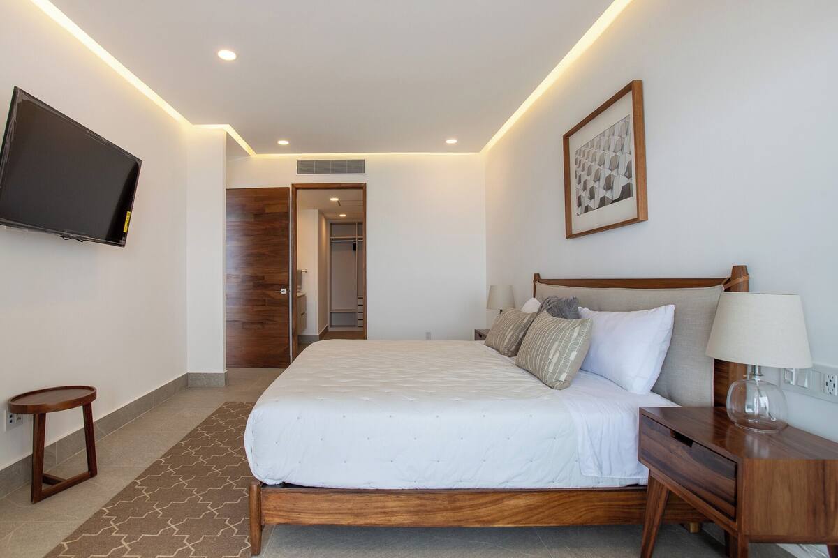 Una habitación de hotel con cama, mesita de noche, lámpara y televisor montado en la pared.
