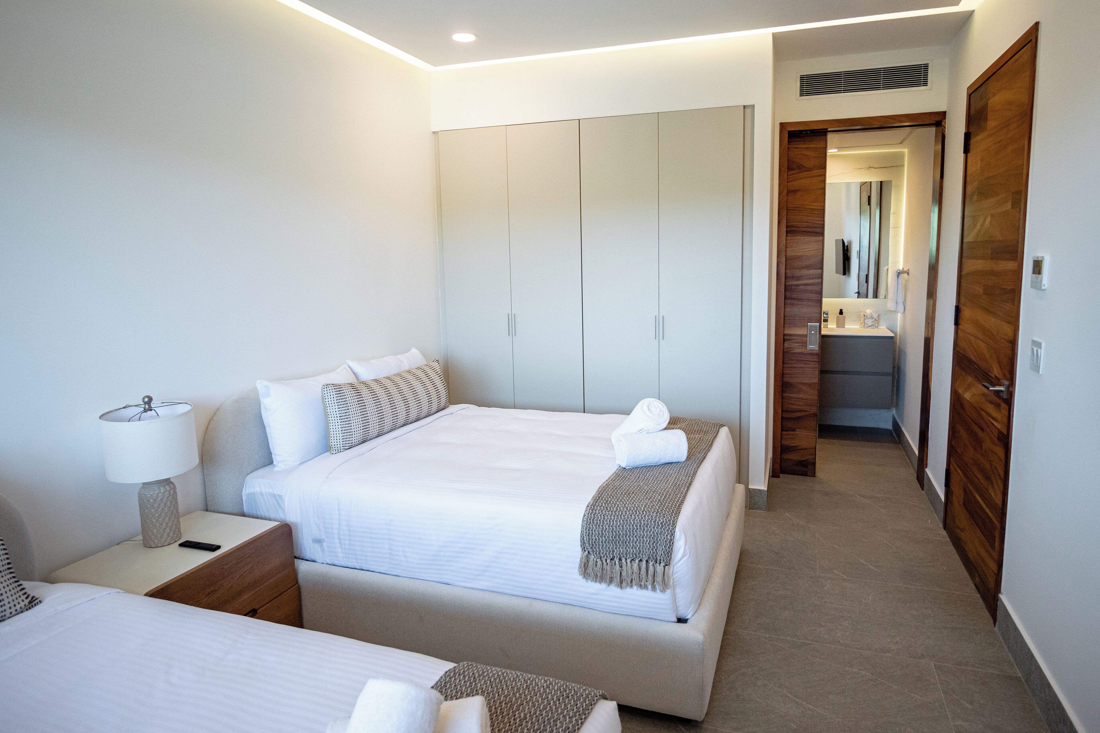 Une chambre d’hôtel moderne, dotée d’un grand lit, d’une table de chevet avec une lampe, d’une armoire en bois et d’une salle de bain visible à travers une porte ouverte.