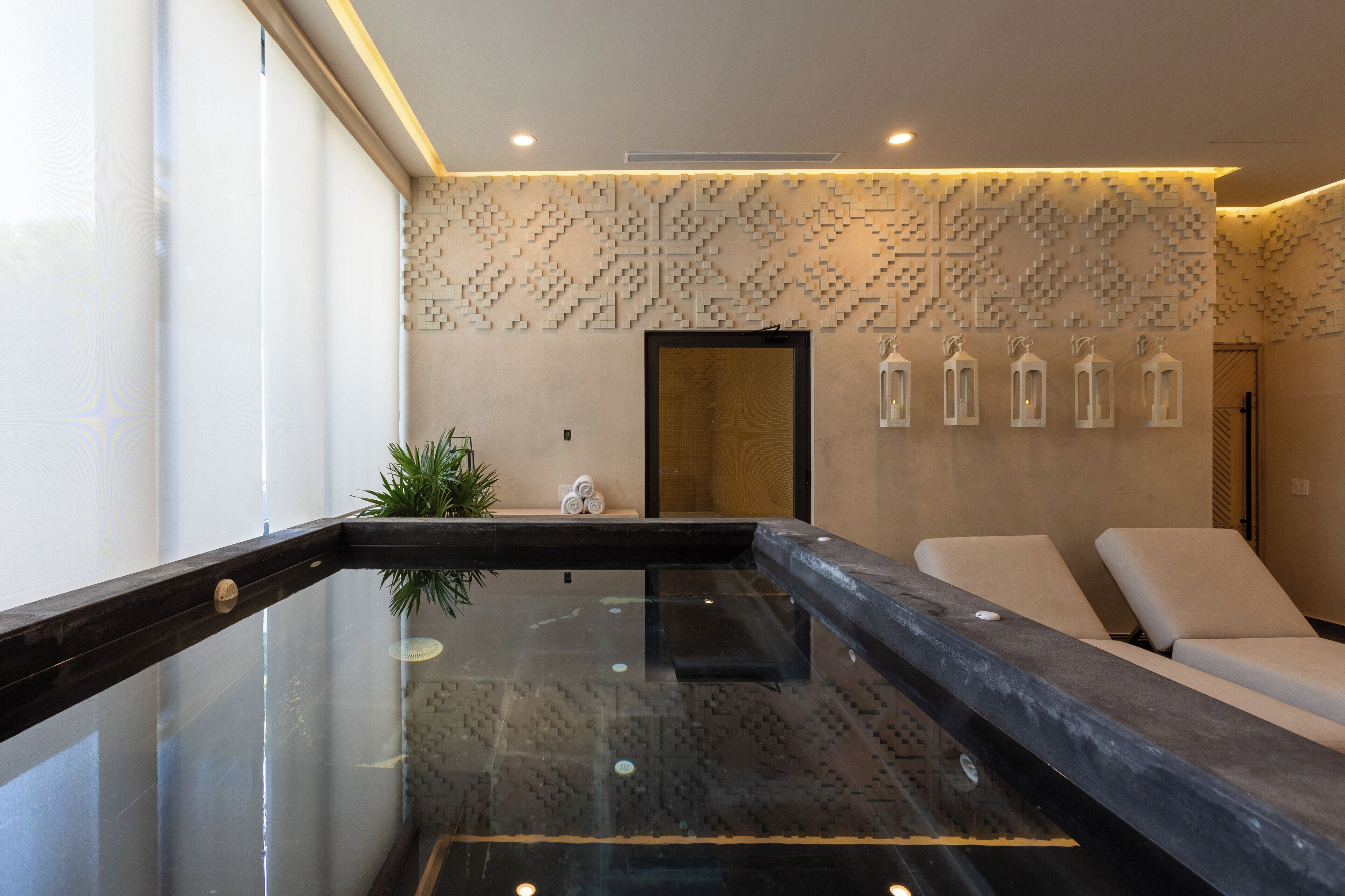 Un área de spa moderna con piscina de vidrio, sillones de descanso y paneles decorativos en la pared.