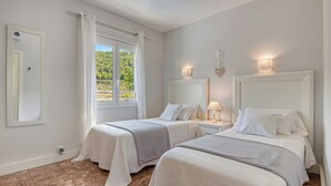 Een hotelkamer met twee bedden, een raam met gordijnen, een wandlamp en uitzicht op groen.