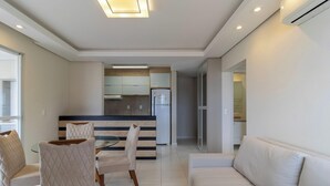 Ein modernes Wohnzimmer mit einer Couch, einem Esstisch mit Stühlen, einer Küchenzeile mit Schränken und einem Kühlschrank, sowie einem Flur, der in einen anderen Raum führt.