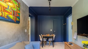 A dining area with a table and chairs, a painting on the wall, a sofa, and a TV on the wall. - Condominium in Floripa 300m From the Beach - CAS (Florianópolis)