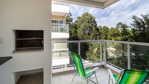 Un balcon avec deux chaises à rayures vertes, une petite étagère et une vue sur les arbres et les bâtiments.