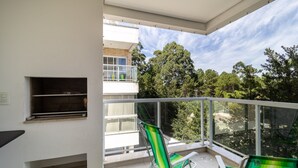 A balcony with two green-striped chairs, a small shelf, and a view of trees and buildings. - Apt w/ Barbecue Near the Beach in Campeche Bve101 (Florianópolis)
