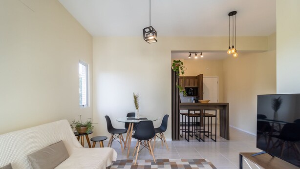 A modern living room with a sofa, a dining table with chairs, a TV, and a kitchen area in the background. - Apt w/ Barbecue Near the Beach in Campeche Bve101 (Florianópolis)