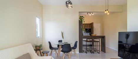 A modern living room with a sofa, a dining table with chairs, a TV, and a kitchen area in the background.