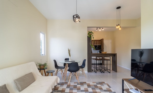 A modern living room with a sofa, a dining table with chairs, a TV, and a kitchen area in the background.