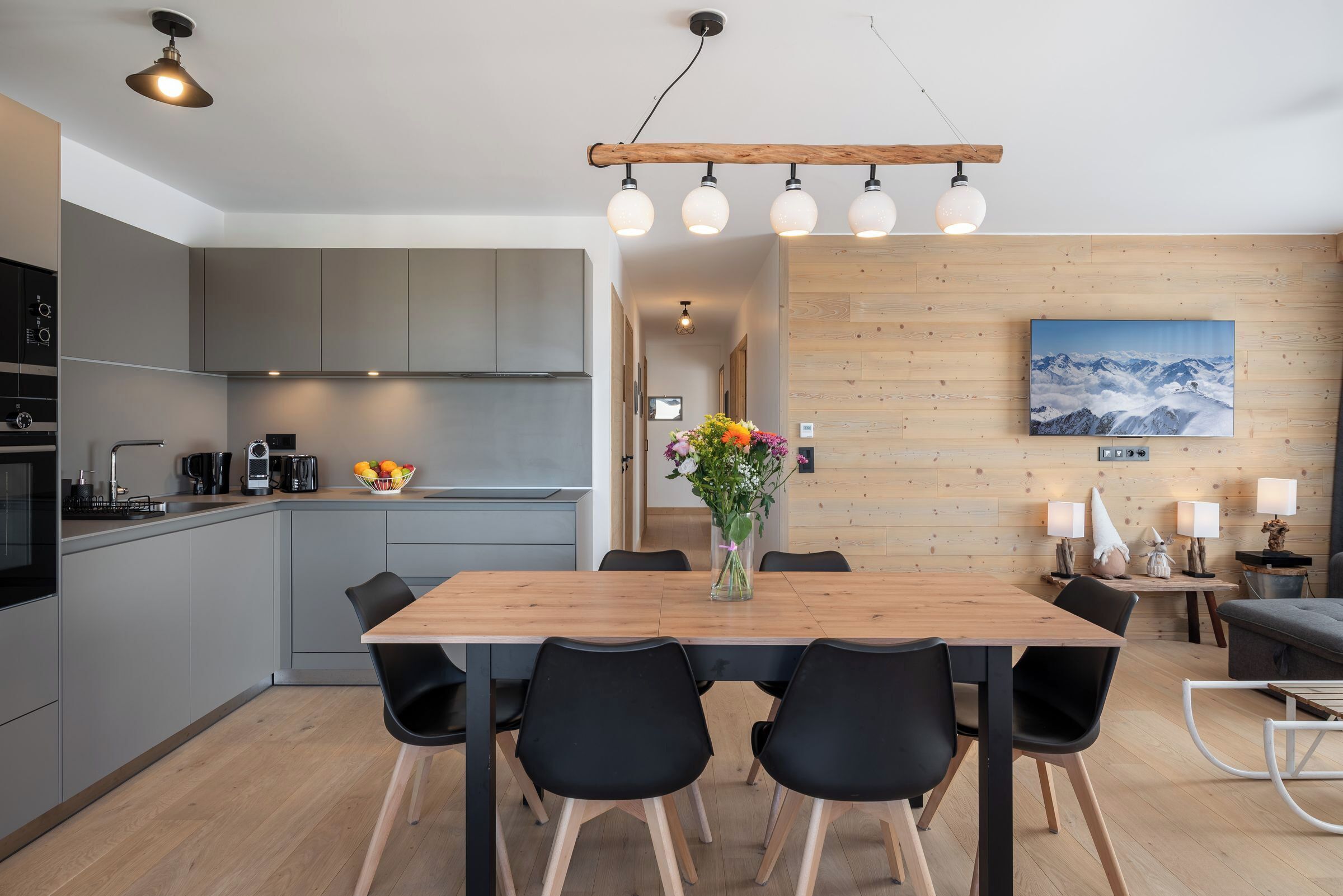 Une cuisine moderne avec des meubles gris, une table à manger en bois et des chaises noires. Un vase rempli de fleurs est posé sur la table.