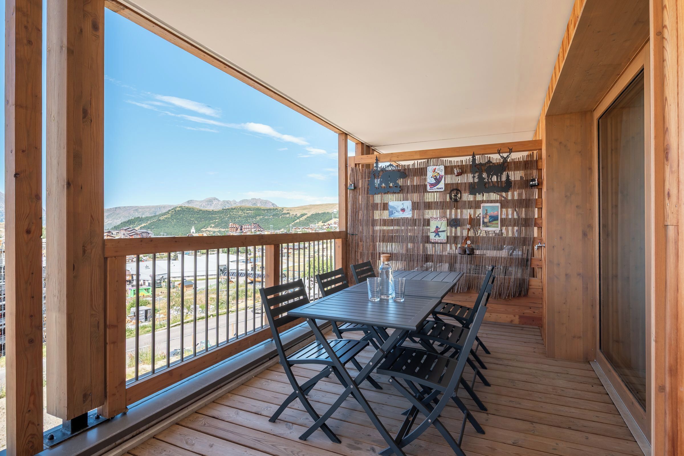 A wooden deck with a table and chairs, a view of mountains, and a bamboo wall with pictures.