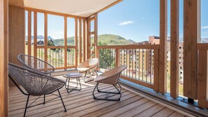 A wooden deck with wicker chairs and a table, overlooking a hilly landscape with buildings in the distance.