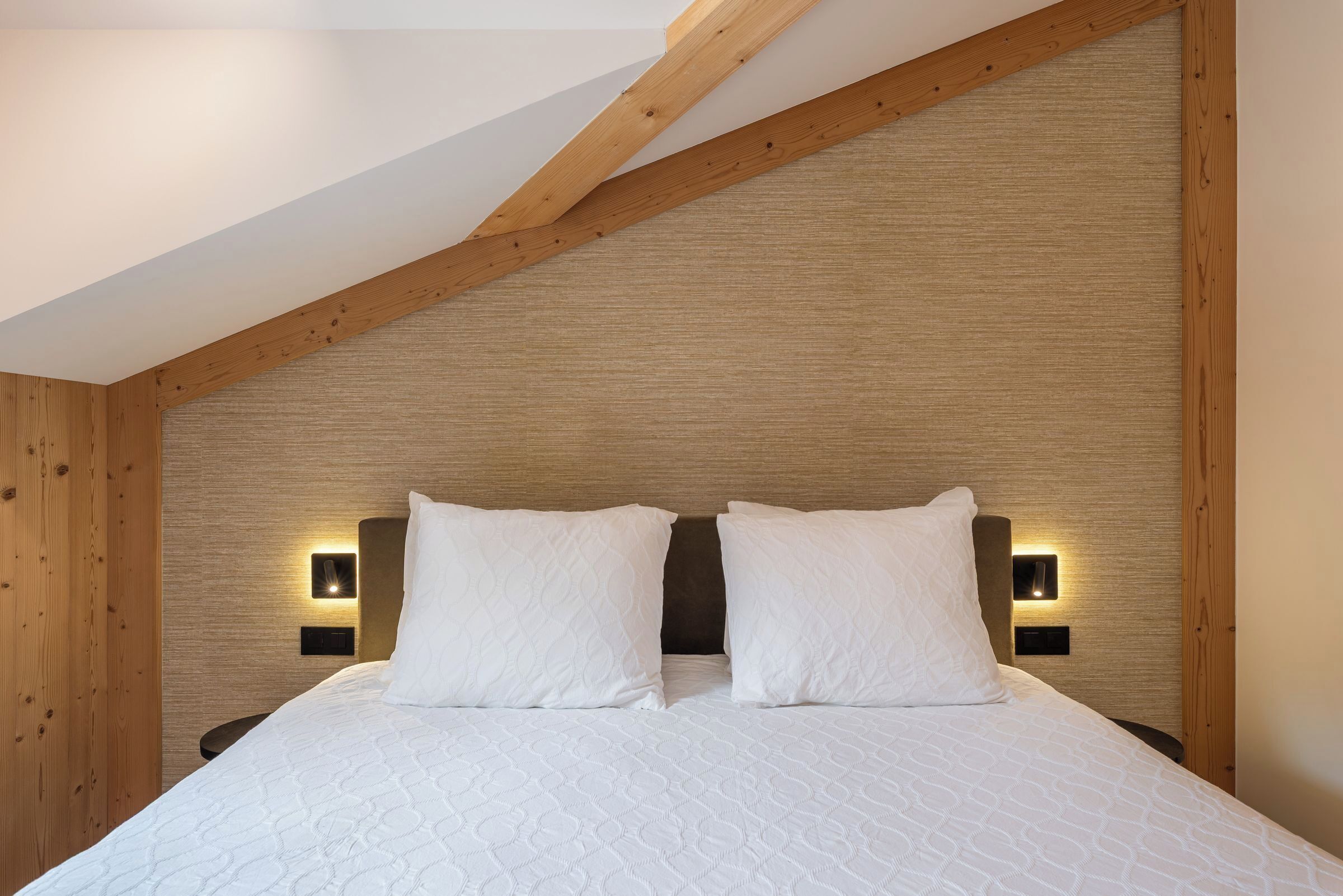 Un dormitorio con techo de madera, una cama con ropa de cama blanca y dos lámparas fijadas a la pared.