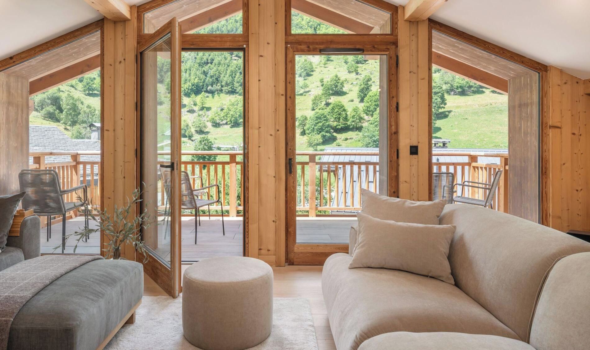 A cozy room with a wooden ceiling, a sofa, and a round ottoman. There is a balcony with a railing and a view of a green landscape.