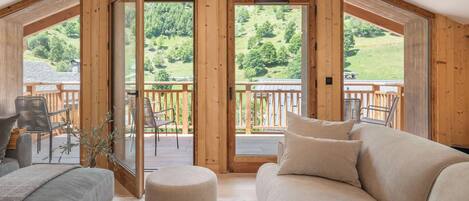 A cozy room with a wooden ceiling, a sofa, and a round ottoman. There is a balcony with a railing and a view of a green landscape.