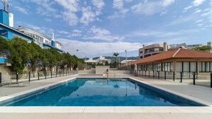 Una piscina con agua cristalina azul, rodeada por un suelo de baldosas y una fila de árboles.