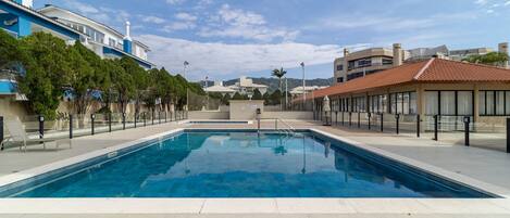 Una piscina con agua cristalina azul, rodeada por un suelo de baldosas y una fila de árboles.