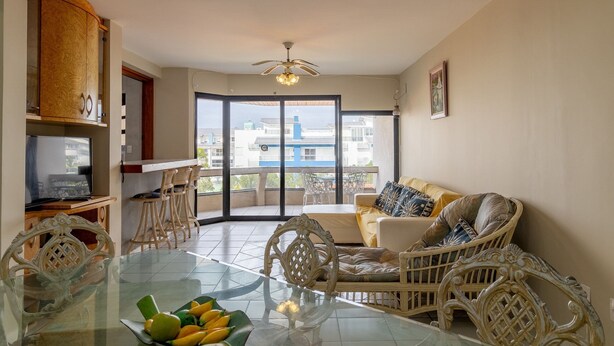 Amplia sala de estar con mesa de comedor de vidrio, mobiliario de mimbre y un balcón con vistas a edificios.