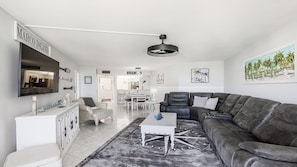 A modern living room with a grey leather sofa, a white coffee table, and a large mirror on the wall. There is a dining area with a white table and chairs, and a kitchen visible in the background. - South Seas Tower II, 1701 (Marco Island)
