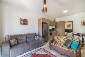 A modern living room with a grey sofa, a wicker sofa, and a kitchen area with wooden cabinets and stainless steel appliances. - Cozy Apartment Just 2 min From the Sea! - Vig0011 (Florianópolis)