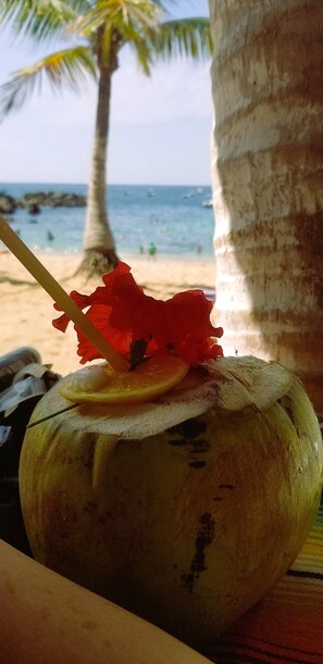 Eine Kokosnuss mit Halm und roter Blume, platziert auf einem Tisch, im Hintergrund ein Strand und Palmen.