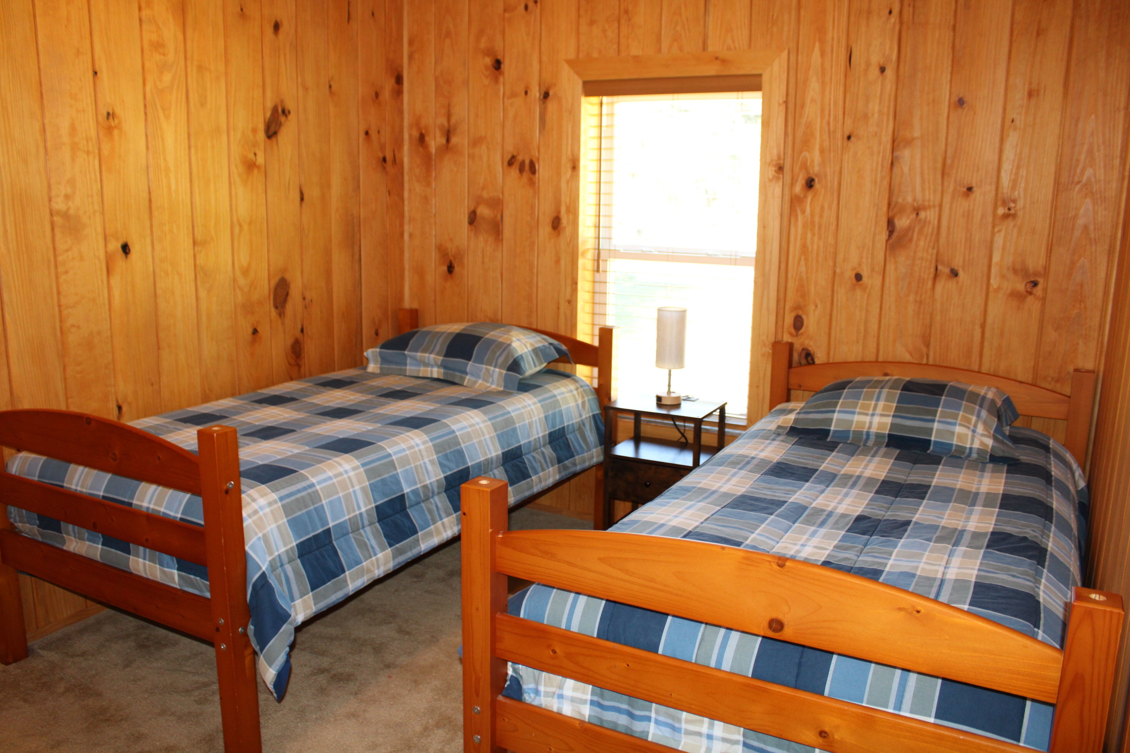 A room with two wooden beds with blue and white checkered bedding, a small wooden table, and a window with blinds.