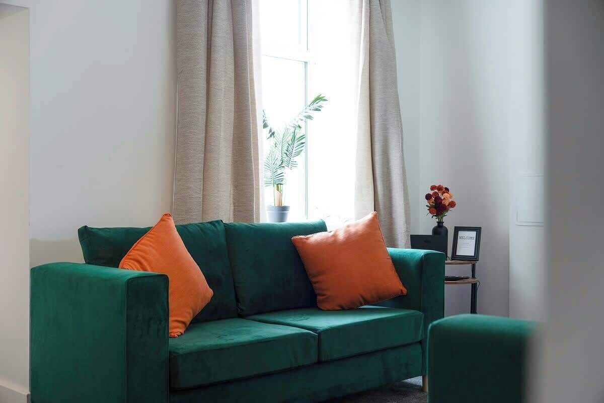 Un canapé vert avec des coussins orange, une plante en pot et une petite table sur laquelle est posé un panneau d’accueil.