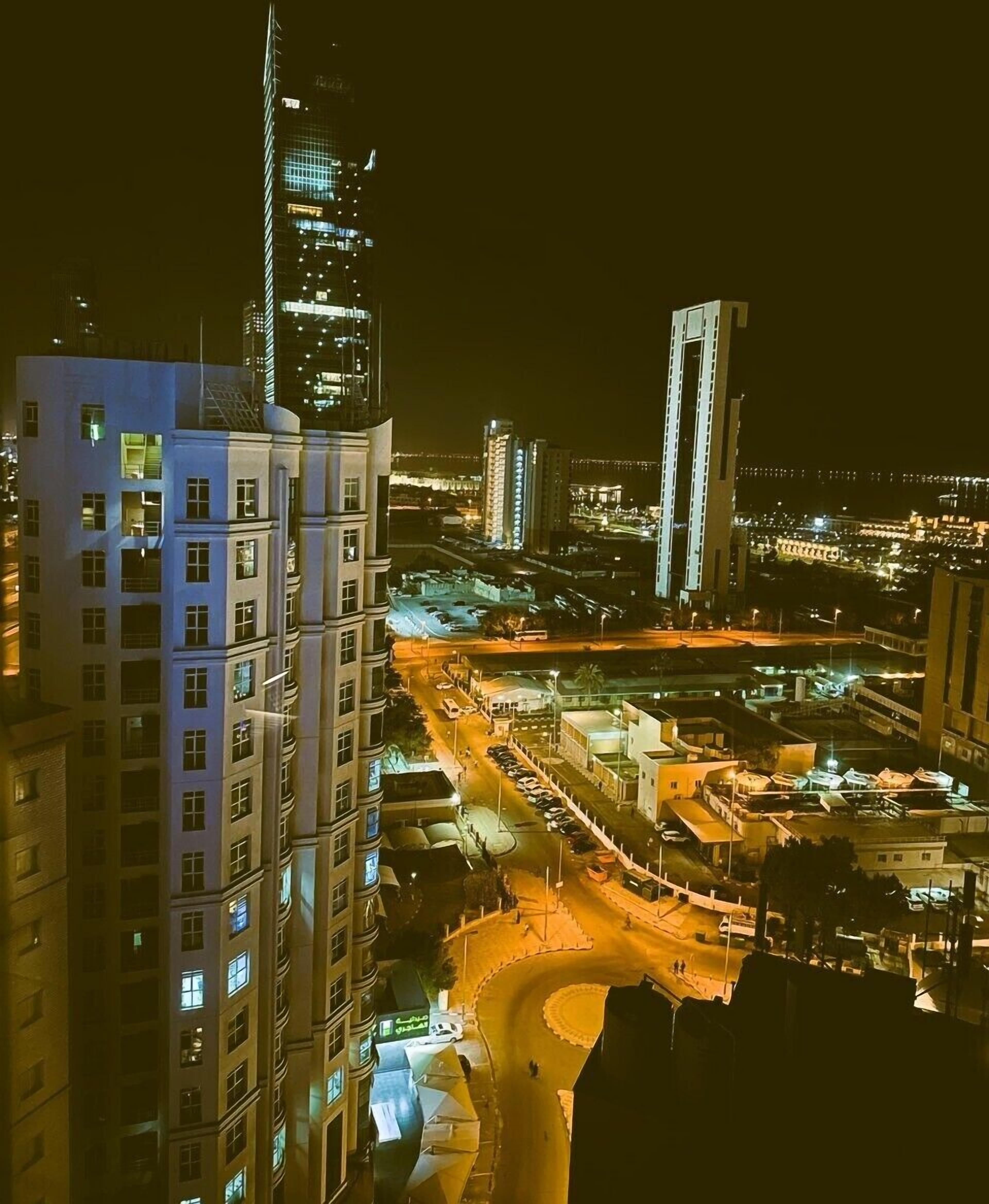 Un paisaje urbano nocturno con edificios altos, una torre destacada y una zona urbana bien iluminada con calles y estacionamiento.