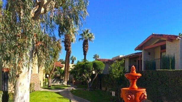 A well-maintained garden with a central orange fountain, surrounded by neatly trimmed grass and palm trees, leading to a building with a red-tiled roof. - Palm Springs 2 bed/2bath condo - Great Location / Resort like setting - 2 pools (Palm Springs)