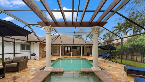 A covered outdoor swimming pool with a pergola, lounge chairs, and a stone-tiled deck.
