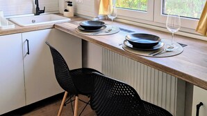 Una cocina moderna con armarios blancos, una encimera de madera y una ventana con cortinas. Hay dos sillas negras y una mesa de comedor con platos y copas de vino.
