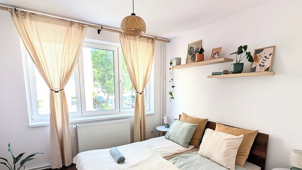 Un dormitorio ordenado con una cama grande, una mesita de noche, una pequeña mesa redonda y un ventanal con cortinas.