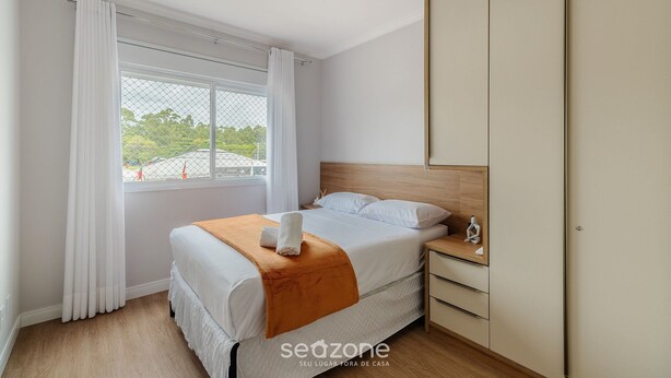 Quarto com cama, mesa de cabeceira, guarda-roupa e janela com cortinas.