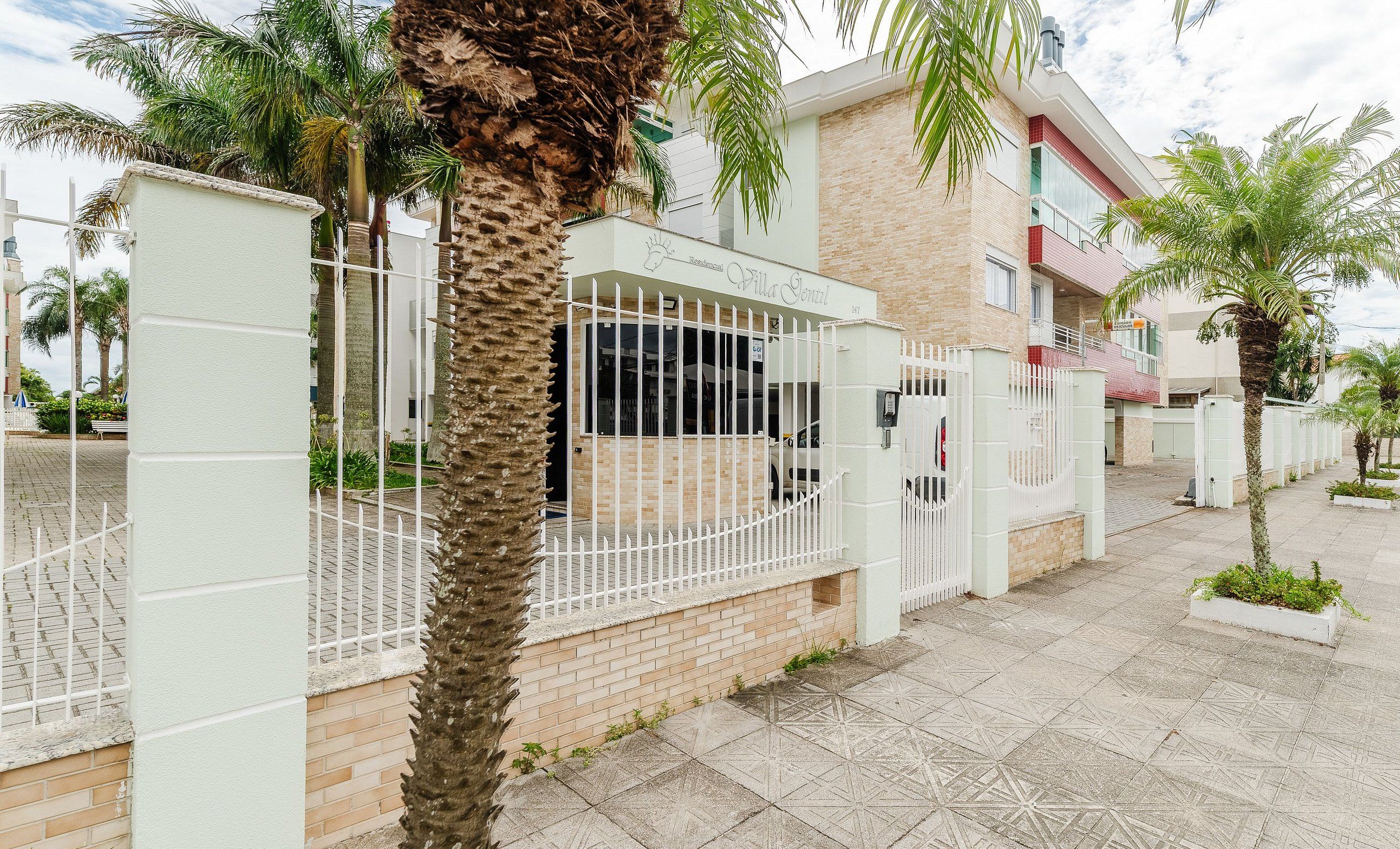 A residential building with a gated entrance, a palm tree in the foreground, and a paved walkway.
