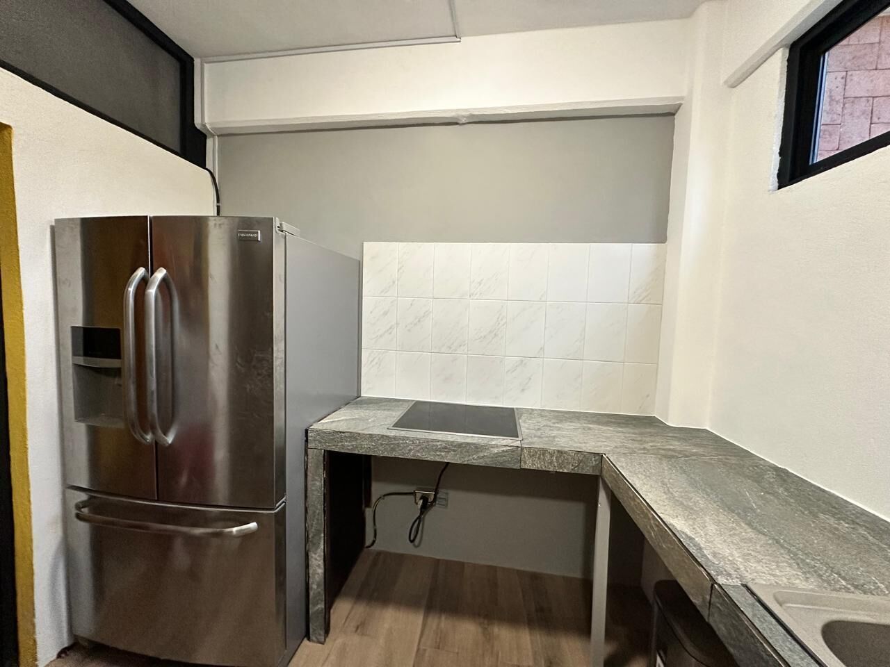 A compact kitchen with a stainless steel refrigerator, a countertop with a built-in stove, and white tiled backsplash.